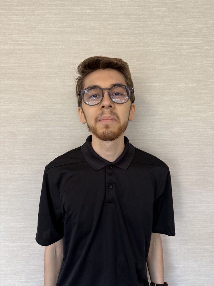 Man wearing glasses and a black polo shirt against a beige wall.
